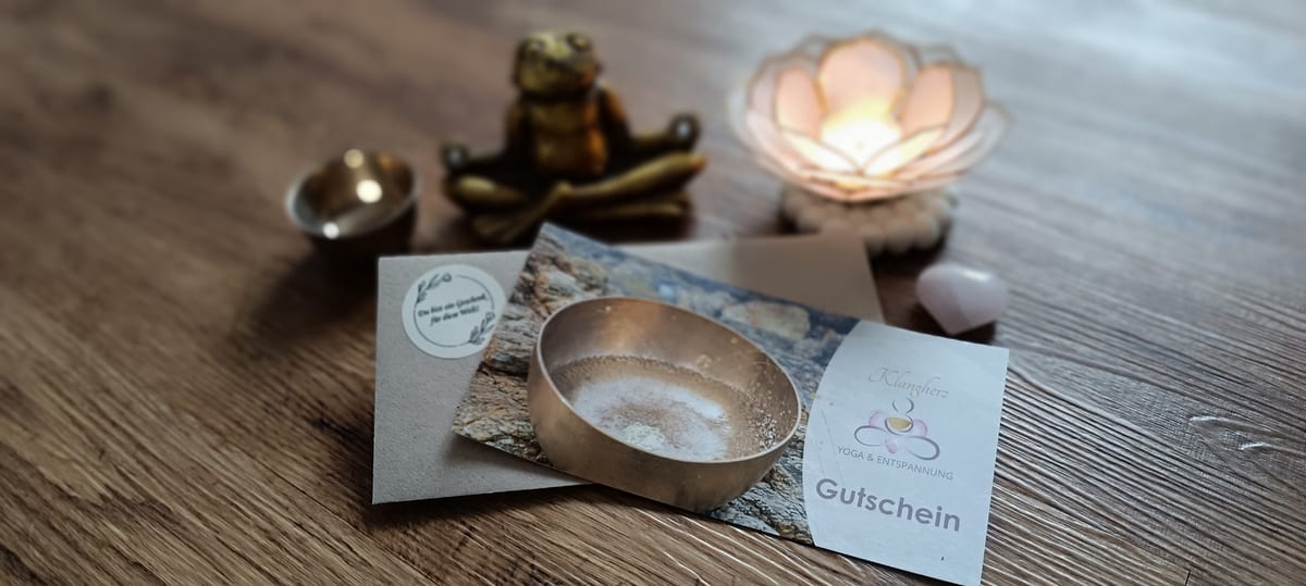Klangherz-Gutschein-Karte mit Kraftpapier-Umschlag und dem Spruch „Du bist ein Geschenk für diese Welt!“, daneben goldener Meditationsfrosch, Lotus-Teelicht und Rosenquarz-Herz auf einem Holztisch