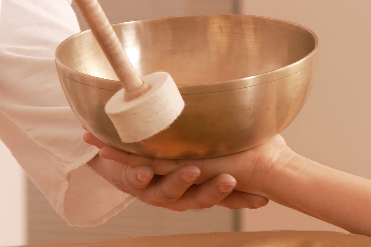 Hände halten eine goldene Klangschale mit Filzklöppel, warme Cream-Rosa-Töne