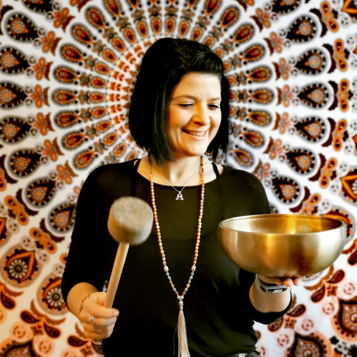 Kerstin Noack lacht mit großer Klangschale und Klöppel in den Händen vor einem Mandala-Tuch