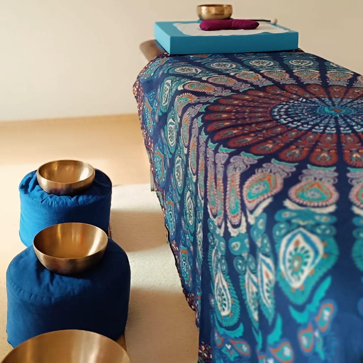 Goldene Klangschalen auf blauen Kissen vor der Massageliege mit Mandala-Tuch