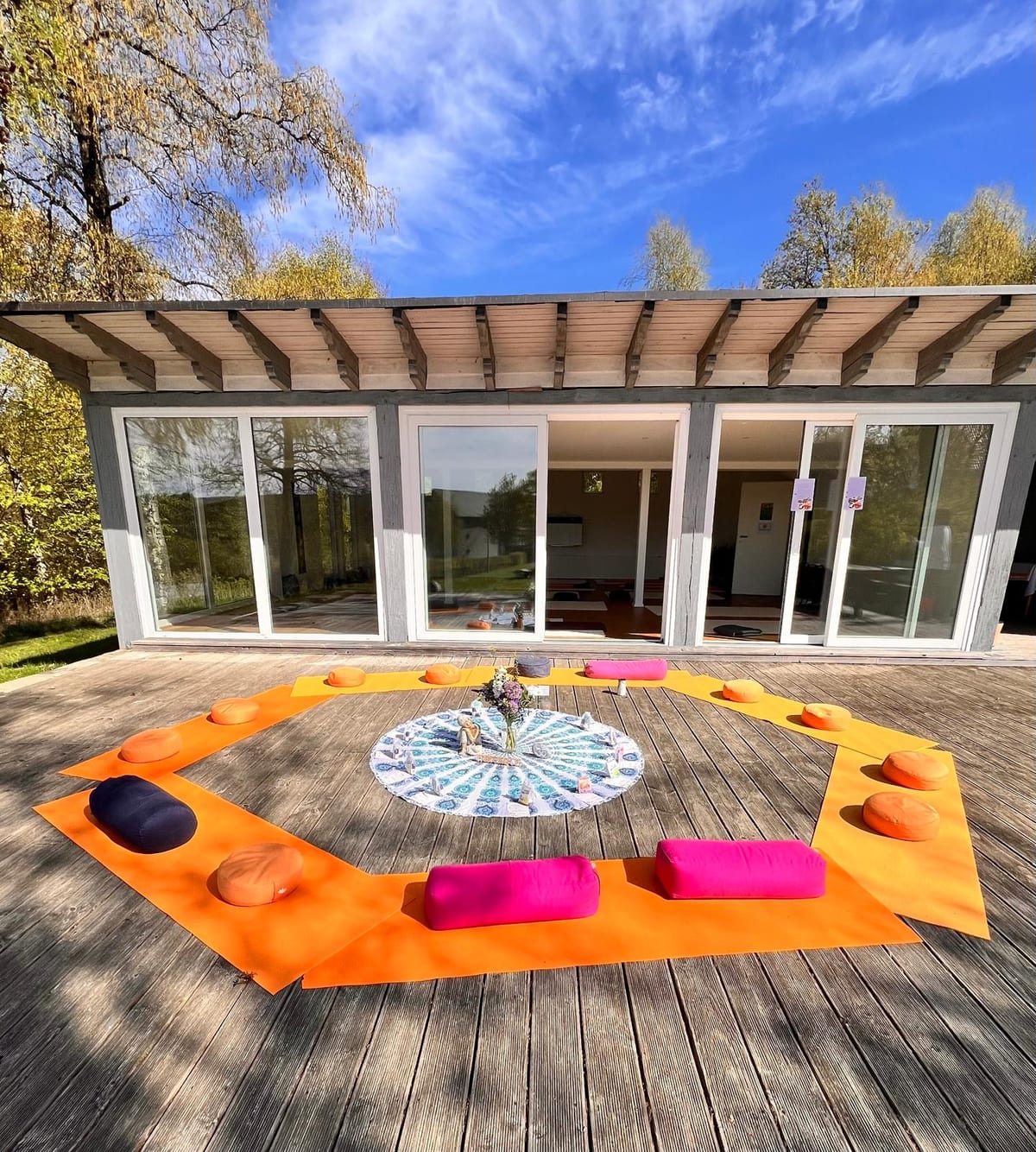 Sonnendeck vor dem Kursraum mit Glasfront: Yogamatten, Bolster und Kissen im Kreis um ein rundes Mandala-Tuch